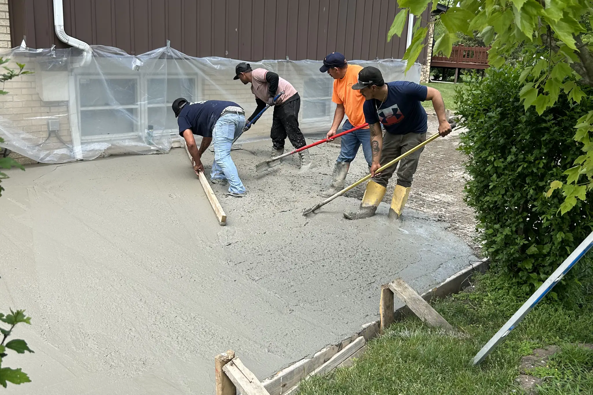 Four workers are smoothing freshly poured concrete on a home site, surrounded by greenery and covered windows Four workers are smoothing freshly poured concrete on a home site, surrounded by greenery and covered windows