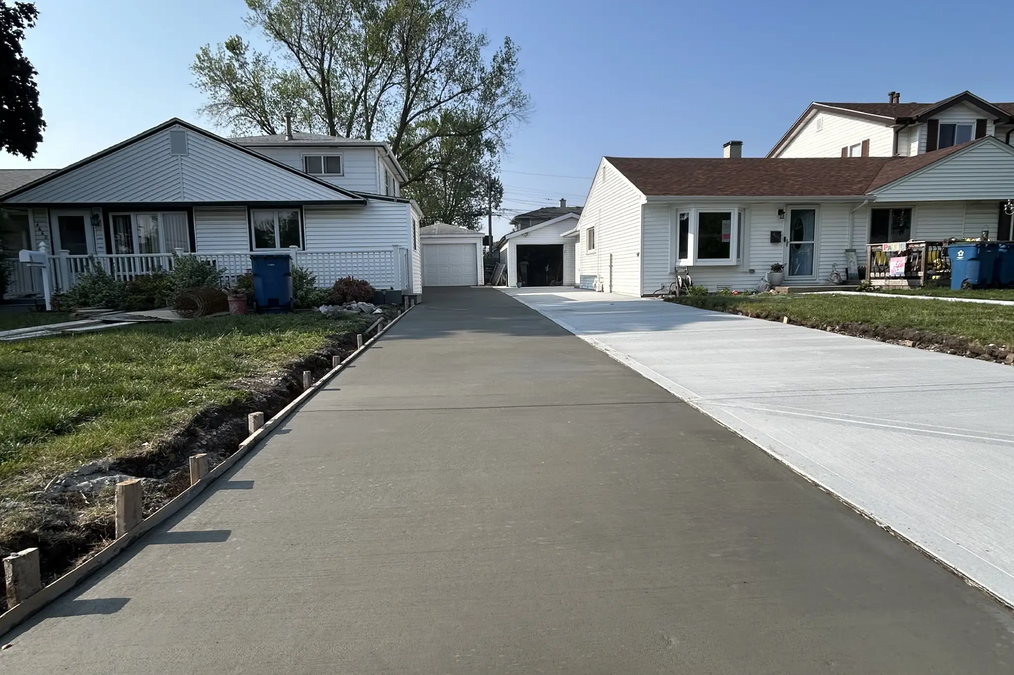 Freshly poured concrete driveway flanked by two houses under a clear blue sky, with wooden forms outlining the driveway edges Freshly poured concrete driveway flanked by two houses under a clear blue sky, with wooden forms outlining the driveway edges