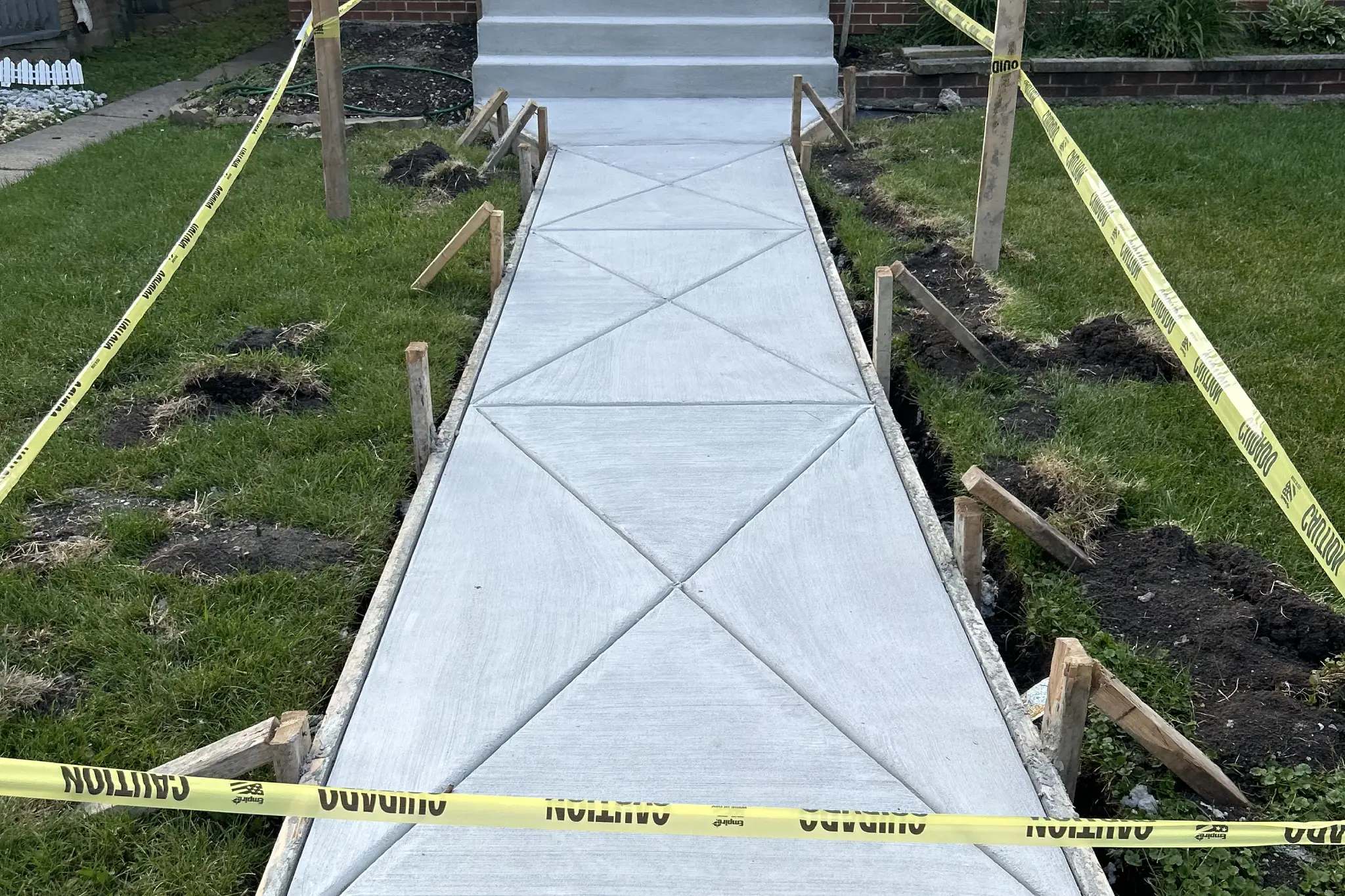 Freshly poured concrete sidewalk leading to a front door, bordered by caution tape and wooden supports, with grass patches on the sides Freshly poured concrete sidewalk leading to a front door, bordered by caution tape and wooden supports, with grass patches on the sides